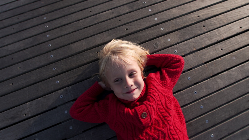 Happy smiling caucasian boy in red sweater is laying on the plank floor outdoor, making funny faces, looking into the camera and laughing. Cool weather | Shutterstock HD Video #1096786507