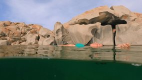 Half underwater scene of woman in blue bikini relaxing floating on sea water of Cala Della Chiesa lagoon with eroded granitic rocks in background of Lavezzi island in Corsica, France. Slow motion