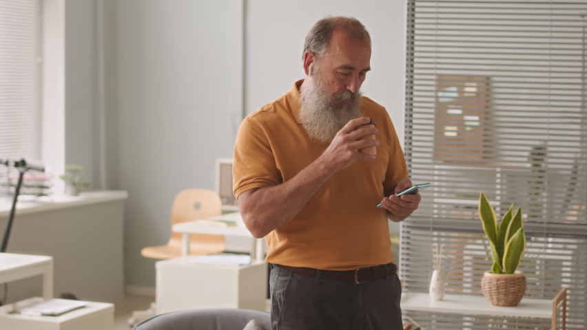 Caucasian old adult man drinking coffee from takeaway cup and listening to music on smartphone in wireless earphones during work in office