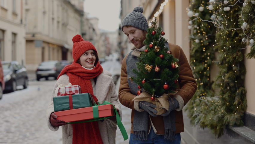The lovely couple is walking, speaking and carrying christmas gifts and tree. Caucasian family is going home from shop, woman holding the gifts and the man is carrying decorated tree