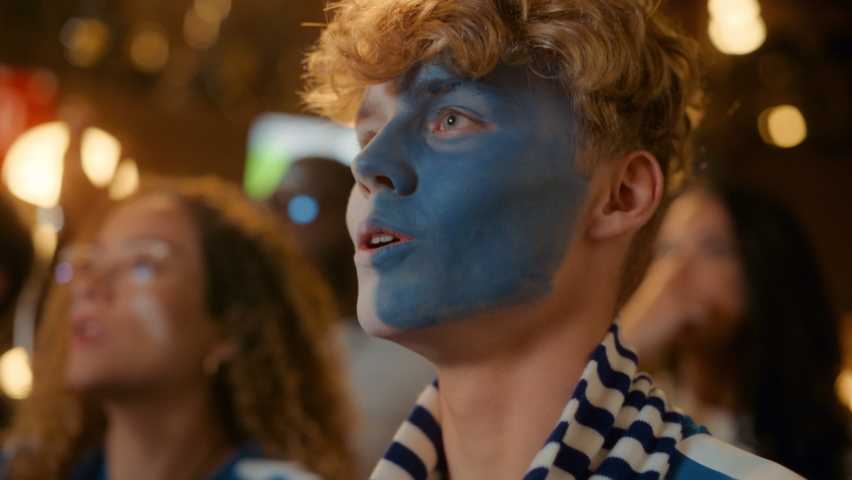 Close Up Portrait of a Handsome Young Soccer Fan with Painted Blue and White Face Standing in a Crowd in a Bar, Chanting, Jumping, Cheering for a Football Team. Friends Celebrate the Goal. Slow Motion
