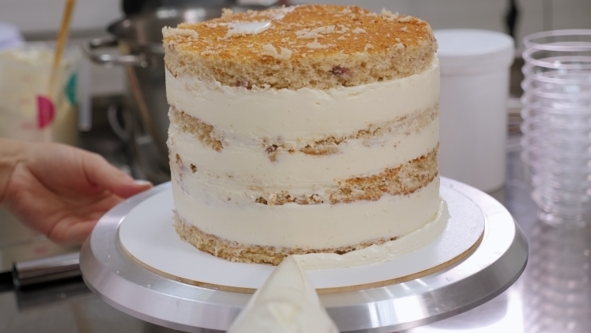 Close-up of a pastry chef rotating a cake on a turntable and squeezing cream from a pastry bag onto the sides of a large cake, the process of making and decorating a cake in a pastry shop.