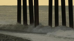 Slow motion video. View of the pier from the side of the sea, the waves break on the stone shore and on the piles of the pier. Cloudy evening.