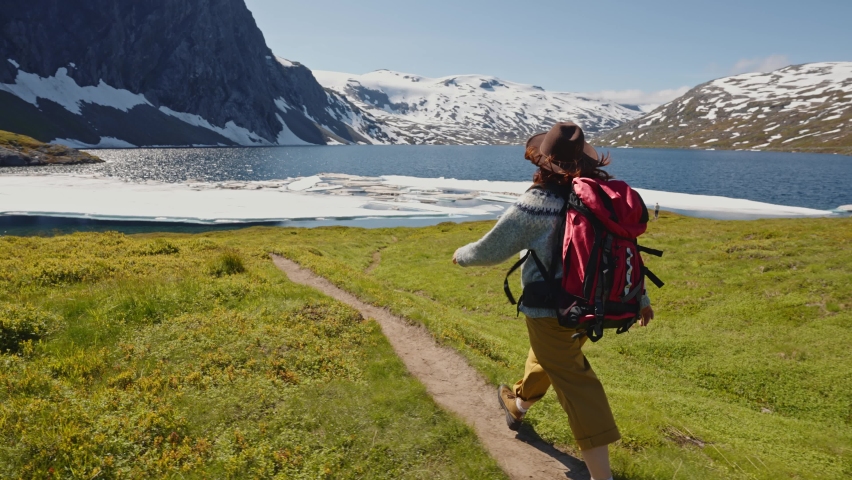 Young woman in a hat with a backpack comes down from the mountains on a background of snow capped mountains