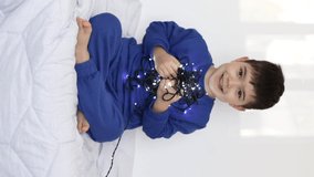 adorable kid preschooler playing with christmas lights inside home bed blanket blue sport suit.child smiling putting light on hand neck untangle have fun.boy hold light close to chest with both hand