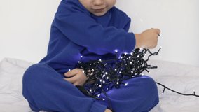adorable kid preschooler playing with christmas lights inside home bed blanket blue sport suit.child smiling putting light on hand neck untangle have fun.boy hold light close to chest with both hand