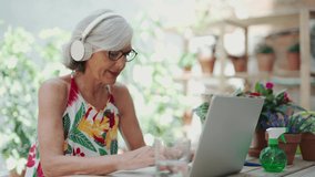 Video of cheerful senior woman listening to music and using her laptop while sitting in greenhouse. - Powered by Shutterstock - Get 15% off with code: PIKWIZARD15