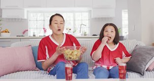 Video of happy asian mother and daughter eating chips and watching match in tv. Family, motherhood, relations and spending quality time together concept digitally generated video.