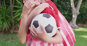 Video of happy asian mother and daughter in soccer shirts embracing in garden. Family, motherhood, relations and spending quality time together concept digitally generated video.