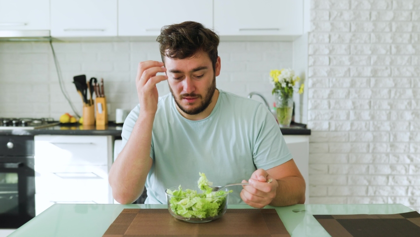 A sad young man looks longingly at a salad, holds a fork with greens in his hand, and a plate of salad in front of him. An exhausting diet haunts, hates healthy food. Stop diet. | Shutterstock HD Video #1096822127