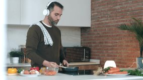 Video of handsome mature man tasting the food he is preparing while listening music with headphones in the kitchen at home.