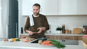 Video of handsome mature man cooking following a recipe from the internet with digital tablet while listening music with headphones in the kitchen at home.