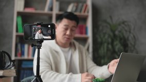 Focus on smartphone screen, pleasant young asian man working on laptop while recording video on his smart phone on tripod. Handsome male freelancer working remotely while staying at home.
