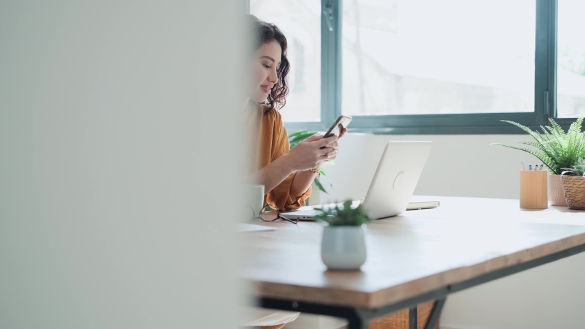 Video of pretty young woman using her mobile phone while working with laptop in the living room at home.