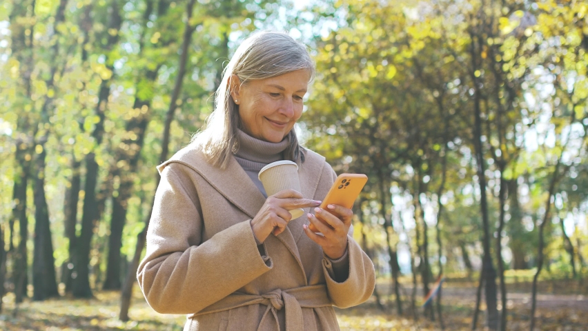 Happy grey-haired beautiful mature woman using smartphone online drinking coffee standing in park in autumn. Attractive cute cheerful female using Internet technologies outside. Smartphone concept. Royalty-Free Stock Footage #1096840851