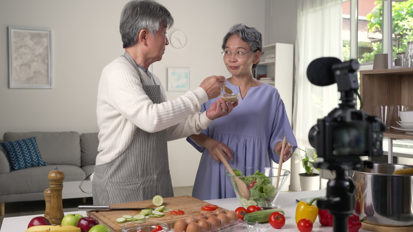 Happy Asian senior couple blogger vlogger and online influencer recording video content on healthy food in the kitchen.
