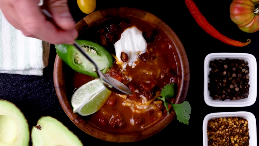 Taking a spoon of chili out of a wooden bowl garnished with lime, green jalapeño pepper, sour cream, cilantro and shredded cheese. Spices and vegetables spilled around it.