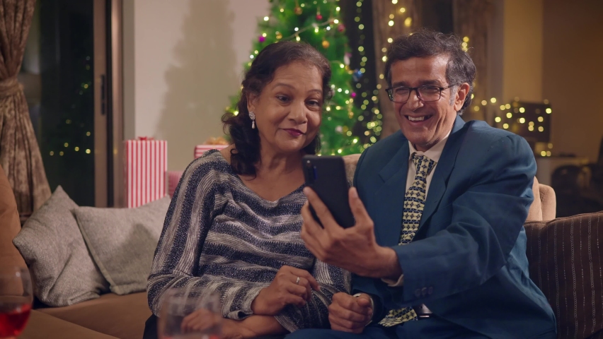 A happy smiling well dressed Indian Asian old or elderly couple or husband and wife sitting together talking to family or friends on a video call using a mobile phone on Christmas or festival holidays Royalty-Free Stock Footage #1096853899