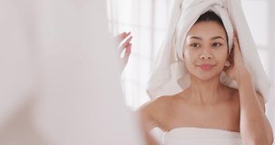 Video of portrait of smiling biracial woman with towel on hair looking in mirror in bathroom. Health and beauty, leisure time, domestic life and lifestyle concept.