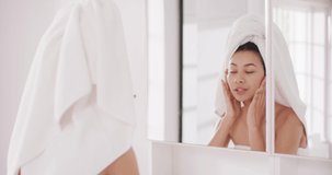 Video of portrait of smiling biracial woman with towel on hair looking in mirror in bathroom. Health and beauty, leisure time, domestic life and lifestyle concept.