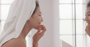 Video of portrait of smiling biracial woman with towel on hair applying cream in bathroom. Health and beauty, leisure time, domestic life and lifestyle concept.