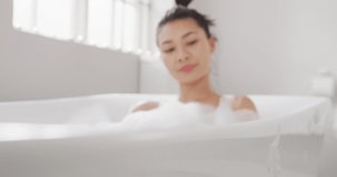 Video of portrait of smiling biracial woman sitting in bathtub in bathroom. Health and beauty, leisure time, domestic life and lifestyle concept.