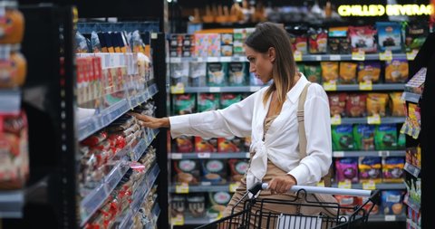 Young Caucasian woman shopper, customer stands in the grocery aisle of a supermarket with a trolley, buys food, reads the composition on the package of spaghetti, is shocked by the prices on the tag, videoclip de stoc