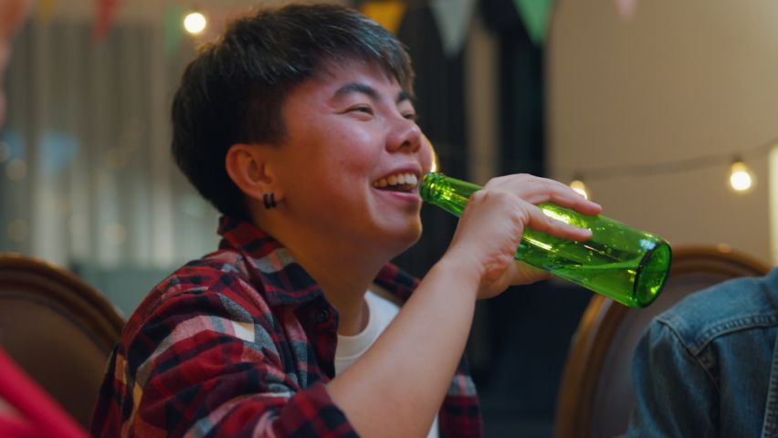 Group of multiethnic adult Asia friend drink alcohol beer bottle glass and fun talk laugh enjoy hangout party at dining table in night life indoor home. Happy hour millennial generation inside house.