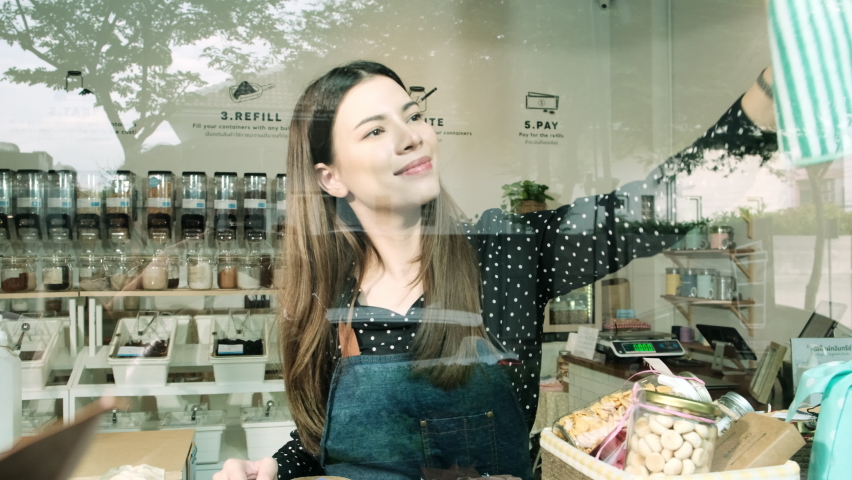 Young Caucasian female shopkeeper works by cleaning glass window displays for shop openings at refill store, zero waste, plastic-free grocery merchandise, and an eco-friendly retail business startup.