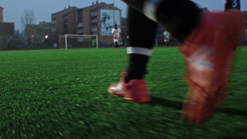 Player walks in football sneakers boots on field during soccer game in small stadium. Teams in cleats shoes are playing friendlie match on green grass in evening. Low angle gimbal close-up shot