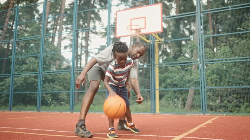 Boy bouncing basketball ball at outdoor court in park. Little kid practicing playing basketball with caring African American father. Sports leisure and weekend. Family time. Basketball game