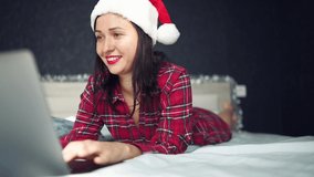 Beautiful excited smiling brunette woman in red pajamas lies in bed in New Year's Santa Claus hat works with laptop in dark interior bedroom. Young female freelancer at home in happy Christmas mood.