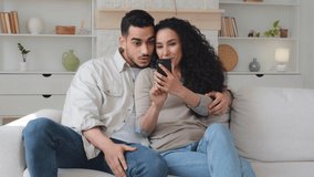 Happy cheerful woman and man Hispanic Latino married couple looking at smartphone smiling talking watching funny video photo sitting on couch at home online fun mobile app using cellphone laughing