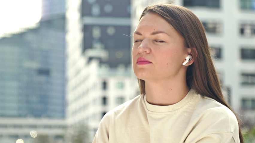 Young attractive woman with wireless headphones in her ears listens to music. Modern European city, summer day. Woman closes her eyes in pleasure.