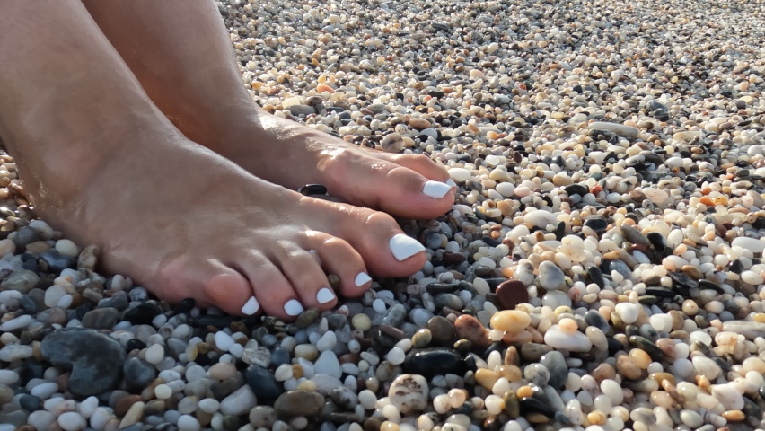 Woman feet with a white manicure on fine gravel is washed by a sea wave with white foam