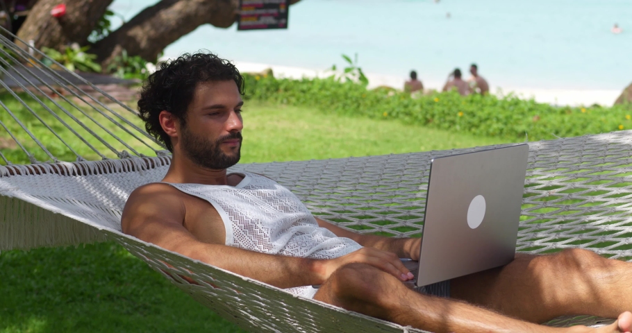 Digital nomad, remote worker, independent location entrepreneur. Successful man works on laptop, he's happy and excited from winning lottery or tender or achieving goals, lying in the beach hammock