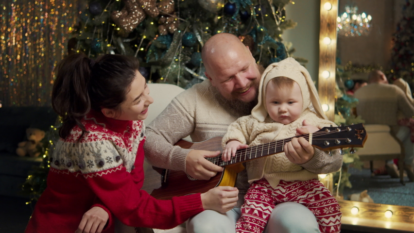 Happy family celebrate christmas eve together at home at decorated background. Mother and father spend time with a cute son teaching to play guitar, listen music. Christmas new year, loving parents Royalty-Free Stock Footage #1096898811