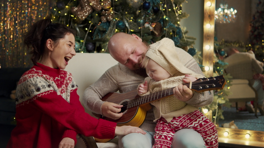 Happy family celebrate christmas eve together at home at decorated background. Mother and father spend time with a cute son teaching to play guitar, listen music. Christmas new year, loving parents | Shutterstock HD Video #1096898811