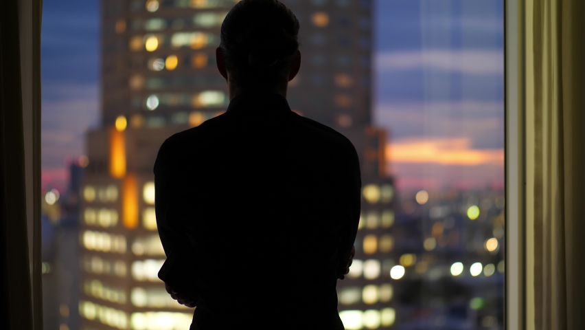 View from back of dark silhouette of a man standing by window, looking out over evening city from room at high floor. Blurred background, tall building with bright windows and sunset sky Royalty-Free Stock Footage #1096909897