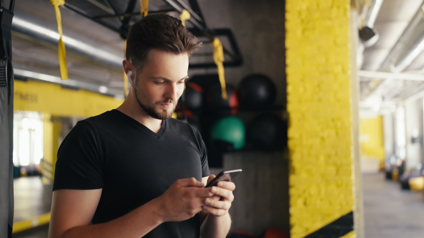 Male fitness influencer in headphones using his smartphone in gym, blogger