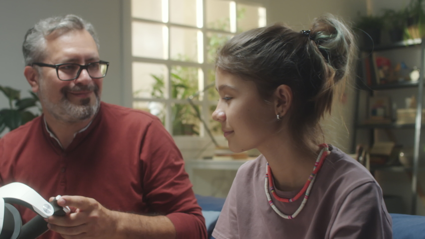 Cheerful father helping teen daughter to put on VR headset while playing game in augmented reality at home