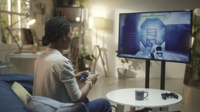 Young African American woman sitting on sofa at home and playing shooter video game on TV