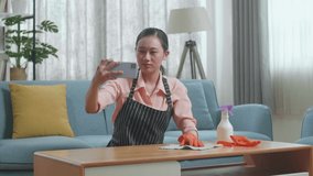 Asian Female Housekeeper With An Apron Having Video Call On Smartphone While Cleaning The Table By The Spray At Home
