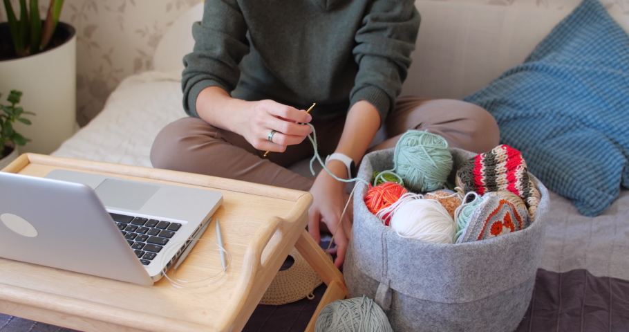 Close-up video of woman learning to crochet. Felt basket with balls of yarn. Anti-stress hobby. Online lessons for amateurs on laptop.