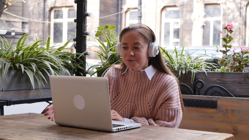 young attractive woman dressed in a pink knitted sweater in a street cafe talking on a video link using a laptop and sitting at a table, wearing white headphones. Video conference dialogue