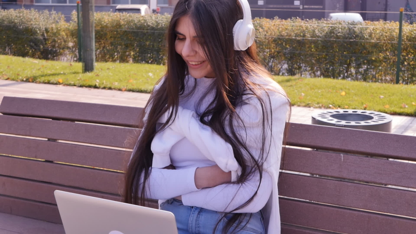 middle eastern pretty young woman with long black hair sitting on park bench using laptop for video call and talking holding computer on her lap and wearing white wireless headphones