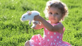 A 2-3-year-old girl plays with a white fluffy rabbit on green grass. Rabbit is a pet. 4k video