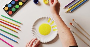 Video of hands painting design on paper plate, with art materials arranged on white background. Art, creativity, homemade, crafts and hobbies concept.