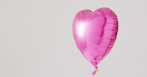Video of shiny pink heart shaped balloon floating on white background, with copy space. Valentine's day, romance, love and celebration concept.