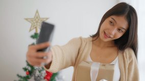 Young asian woman having fun taking light cheerful selfie and video call talking with her friend. Female showing gift box while on video conversation with friend. Happy new year, Thanksgiving